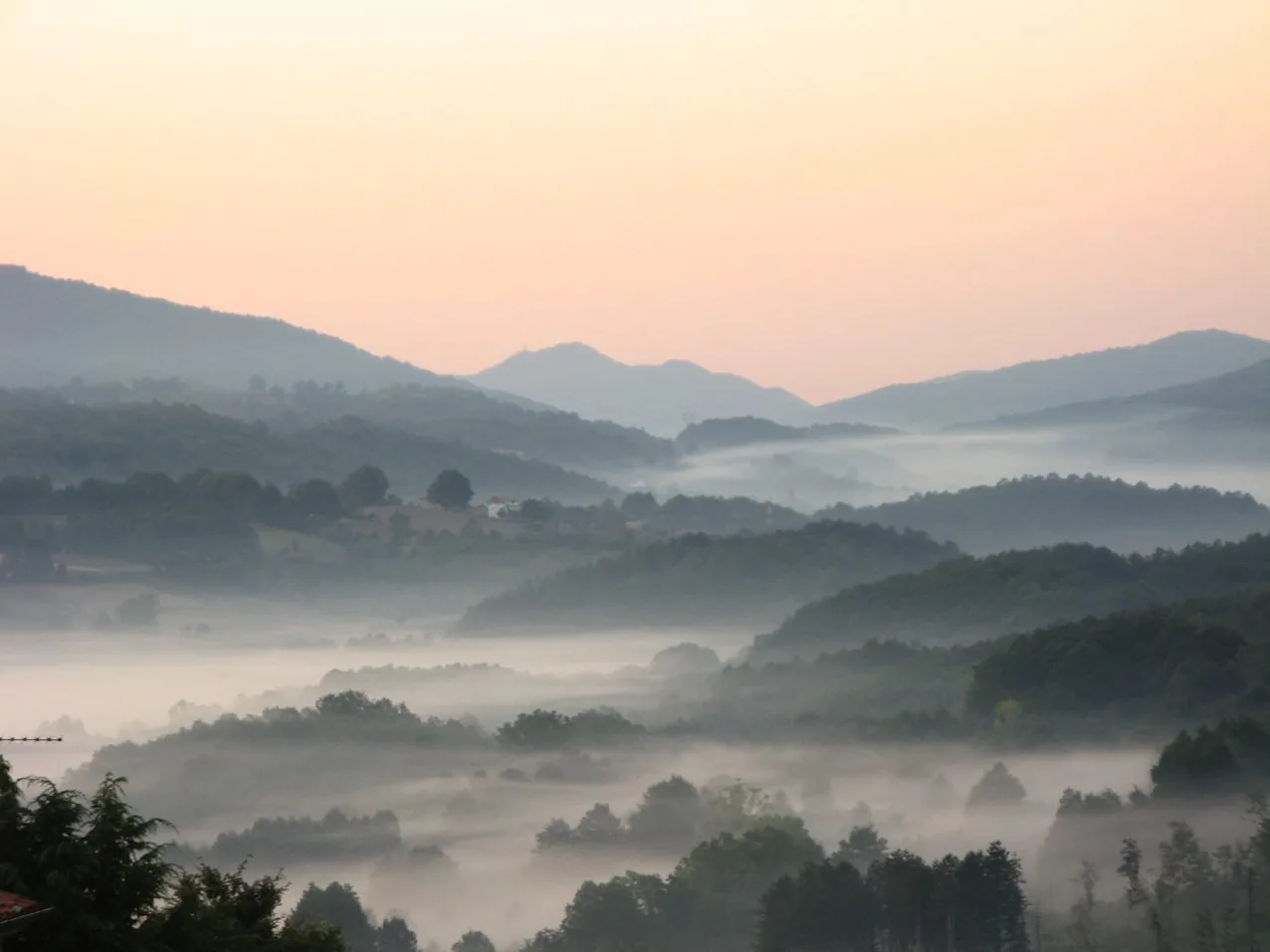 La valle del fiume Amato all'alba, con la nebbia tra le colline boscose e i profili delle montagne contro il cielo rosato